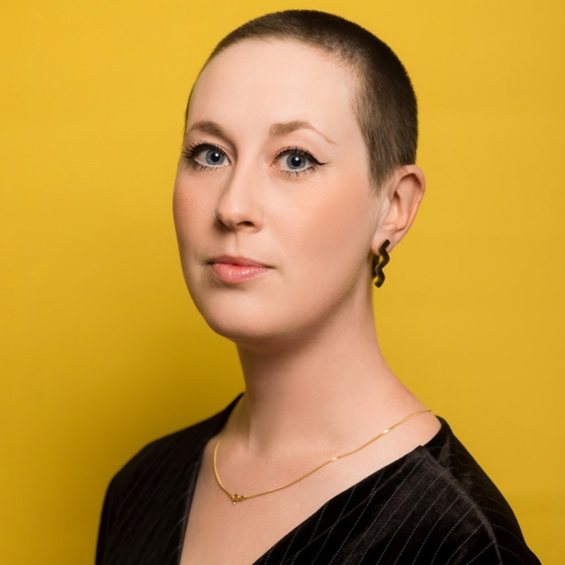 Zoë Programme Manager at Pie Factory Music. Woman with short close shaved hair, with a black zig zag earring and wearing a black top. Yellow background