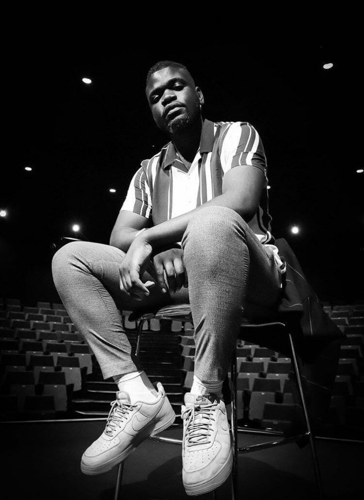Emerging Artist Elijah Femi. Elijah is a young man sitting on a stool in a performing venue looking at the camera