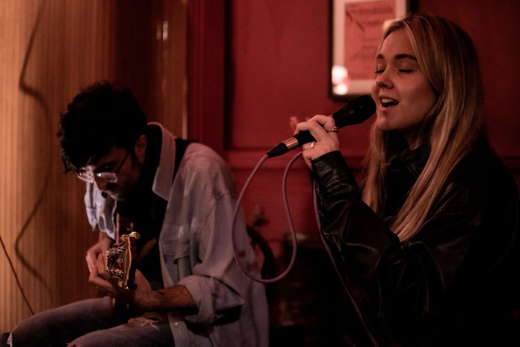 Emerging Artists Maxine and Oren performing at Wantsum Music gig. Maxine is a young woman with blonde hair singing into a microphone. Oren is a young man in a denim shirt playing guitar.