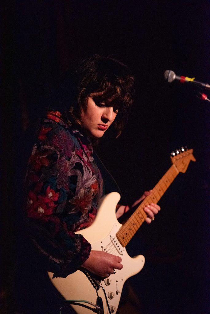 Emerging Artist Claire a young woman in floral blouse playing the guitar