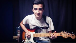 Young man with dark hair wearing white t shirt playing guitar