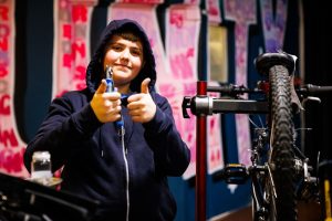 A young man wearing a blue hoodie standing next to a bike with a tool in his hands and making the thumbs up sign
