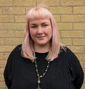 Daisy Operations Manager at Pie Factory Music. Woman with shoulder length pale pink hair and a fringe wearing a black top and long necklace. Yellow background