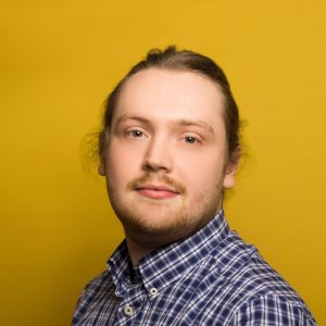 Adam Programme Assistant at Pie Factory Music. Blonde haird man with short blonde beard and long hair in a ponytal wearing a blue and white checked shirt. Yellow background