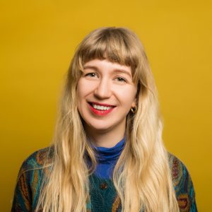 Moa Creative Practitioner at Pie Factory Music. Woman with long blonde hair and a fringe, wearing red lipstick and a blue, green and orange jumper. Yellow background
