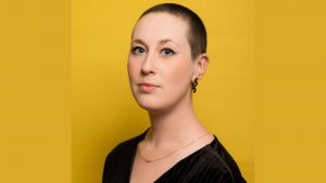 Zoë Programme Manager at Pie Factory Music. Woman with short close shaved hair, with a black zig zag earring and wearing a black top. Yellow background