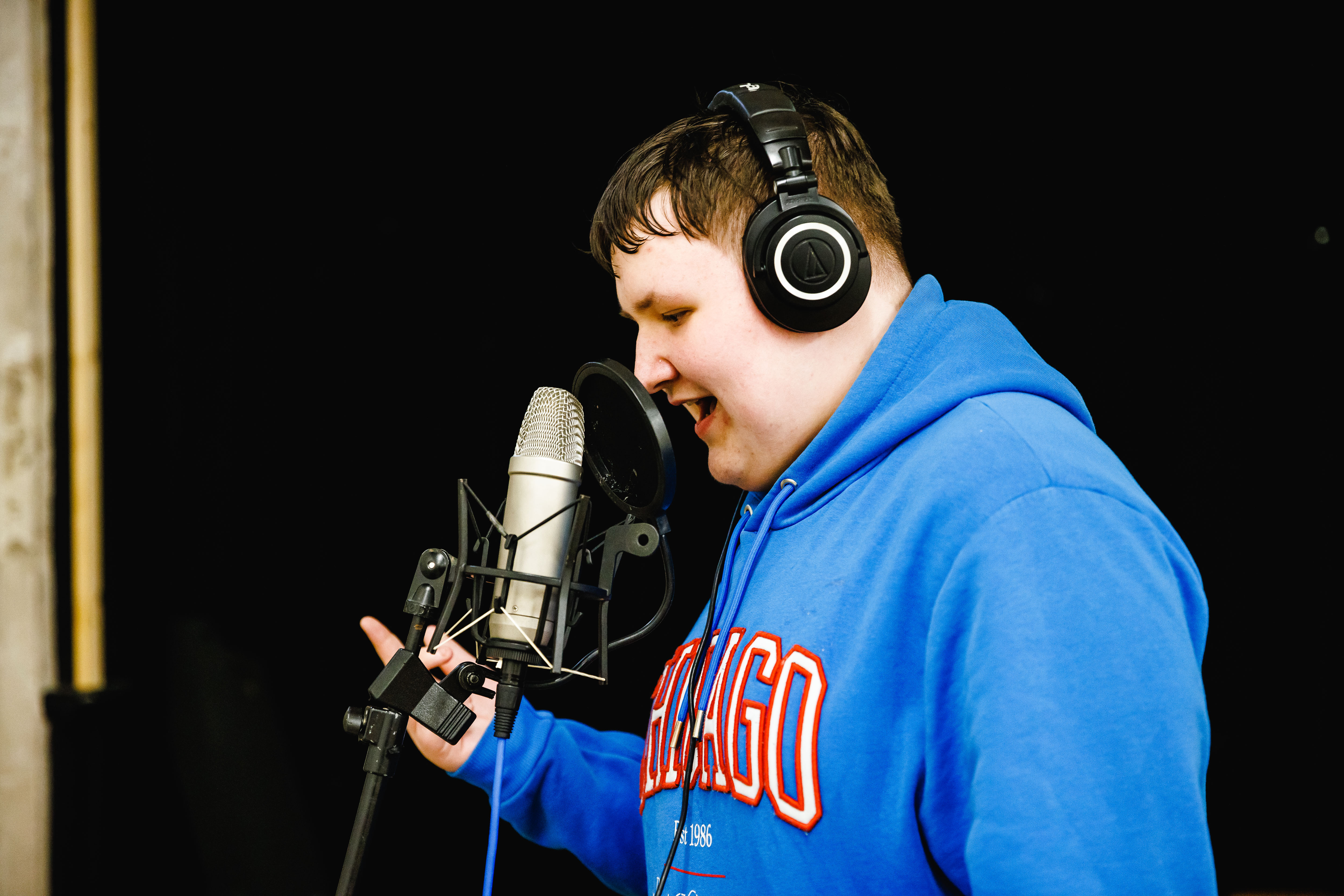 Young person in blue hoodie in front of a mic wearing headphones singing