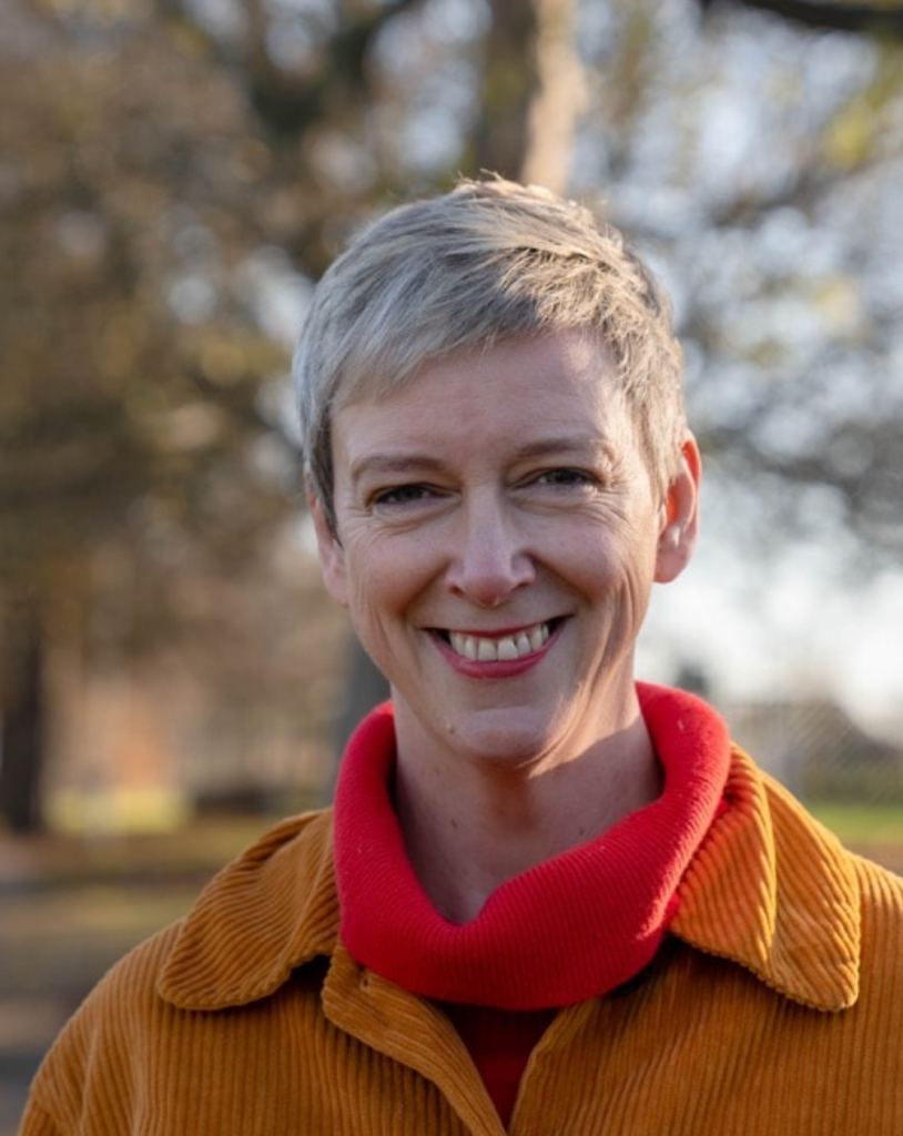 Polly Billington MP - a white woman with short grey-blonde hair with red lipstick smiling. She wears a red roll neck jumper under an ochre courderoy jacket.