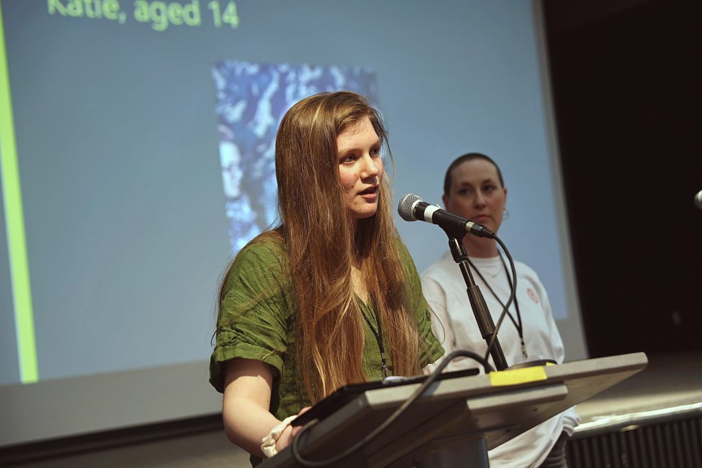 A young woman speaks into a microphone at an event, with another person standing behind her.