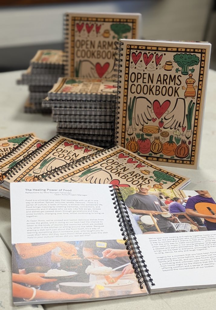 A stack of spiral-bound “Open Arms Cookbook” books with illustrated covers featuring hearts, vegetables, and open hands. Two copies are open, showing colourful pages and photos of people cooking together.