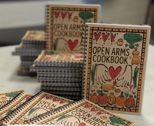 A stack of spiral-bound “Open Arms Cookbook” books with illustrated covers featuring hearts, vegetables, and open hands. Two copies are open, showing colourful pages and photos of people cooking together.